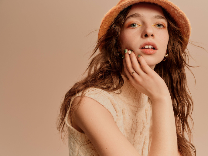Portrait of a young woman with long wavy brown hair, wearing a hat and a cream sweater, posing in front of a peach background.