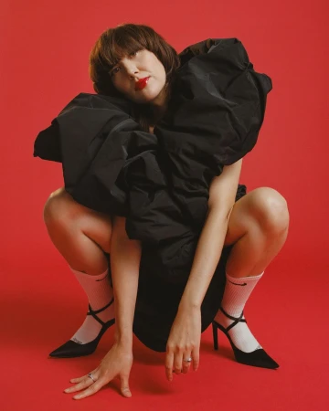 Woman wearing voluminous black puff dress, white socks and black heels crouching against vibrant red backdrop