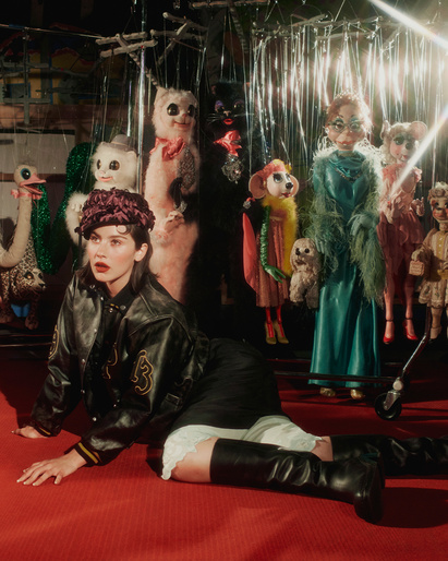 Woman in varsity jacket poses on red carpet with vintage puppets. Fashion editorial with unique set design and retro styling.