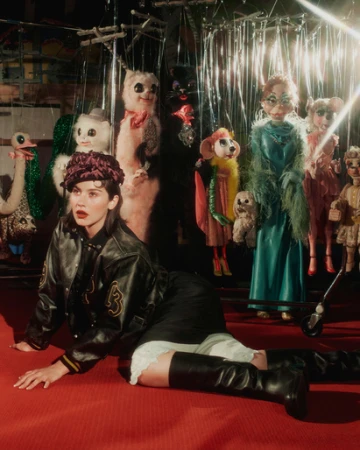 Woman in varsity jacket poses on red carpet with vintage puppets. Fashion editorial with unique set design and retro styling.