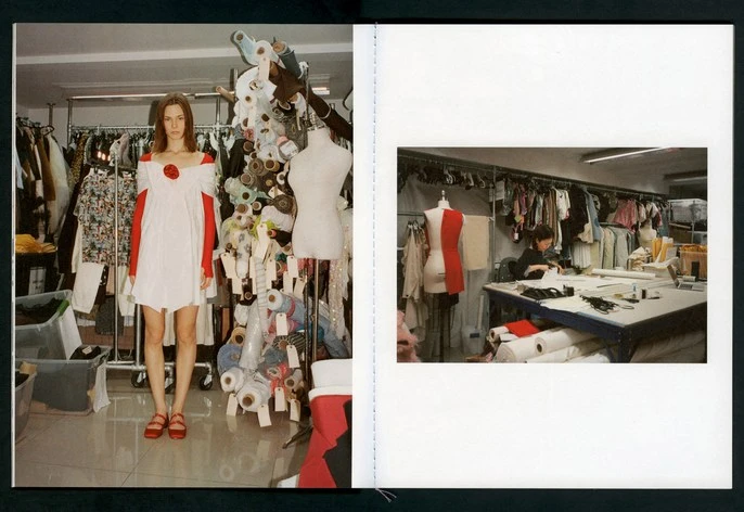 A fashion shot in a dressmaker's atelier with a slender female model and a seamstress at work amidst racks of clothing and fabric rolls.
