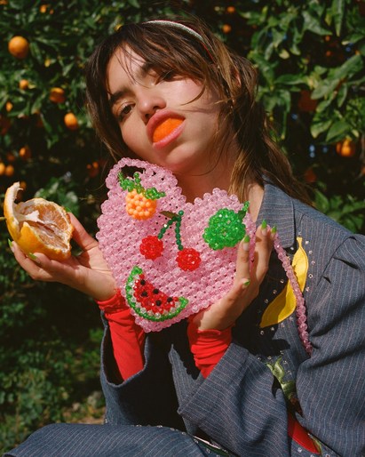 Young woman with bangs holds a beaded heart purse while sitting outdoors amongst orange trees on a sunny day with a playful pose.