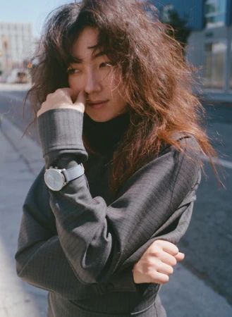 Portrait of an adult woman with long brown hair wearing a grey turtleneck sweater with a watch on a city street with natural light.
