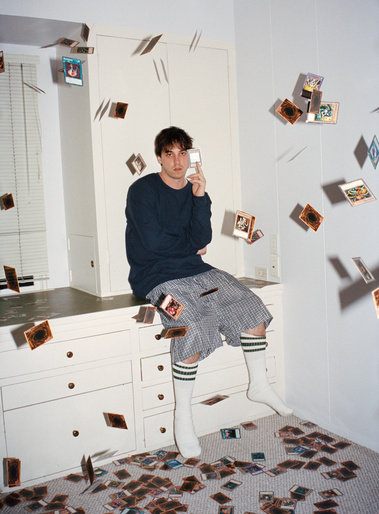 Young adult male sitting on a dresser surrounded by scattered Yu-Gi-Oh cards in a room with white walls and a neutral color palette.