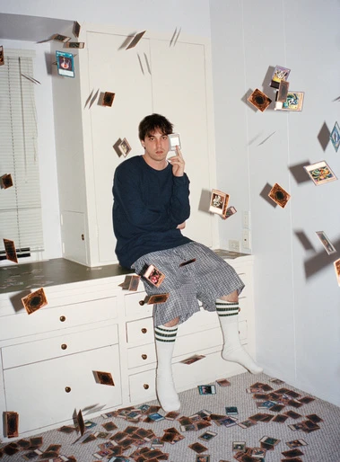 Young adult male sitting on a dresser surrounded by scattered Yu-Gi-Oh cards in a room with white walls and a neutral color palette.