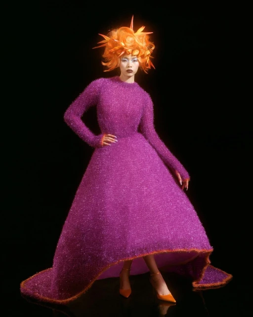 Woman in a bright magenta gown with orange trim and matching spiky orange hair posing on a black background in a fashion editorial shot.