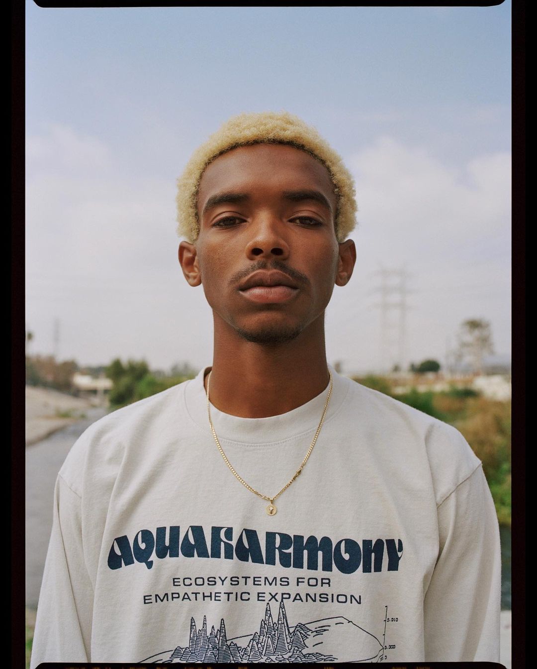Portrait of a young adult male with bleached hair wearing a graphic t-shirt in an outdoor setting with a neutral expression.