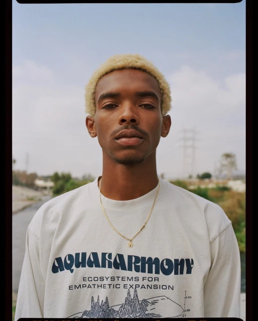 Portrait of a young adult male with bleached hair wearing a graphic t-shirt in an outdoor setting with a neutral expression.
