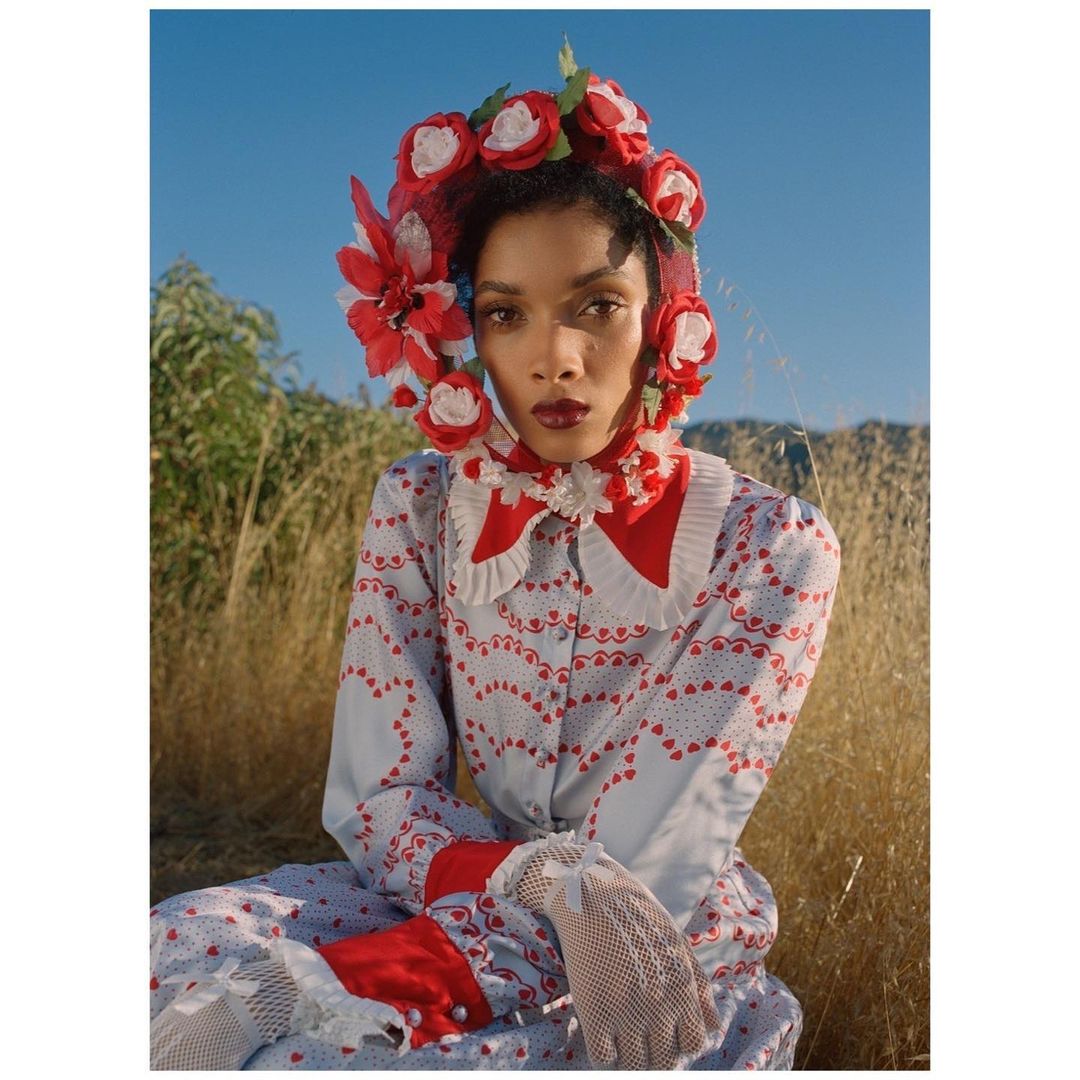 Woman in vintage dress with flower headpiece poses outdoors in grassy field under blue sky; fashion, portrait, editorial photography.