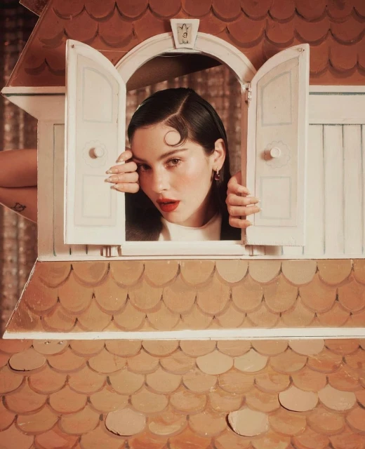 A young woman with dark hair and red lipstick peers through the open shutters of a gingerbread house, captured in a close-up portrait.