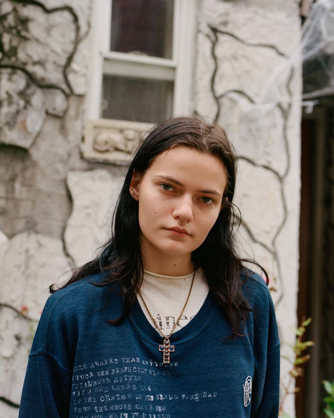 Young woman with dark hair wearing a printed blue sweater and cross necklace posing in front of a stone wall on a cloudy day.