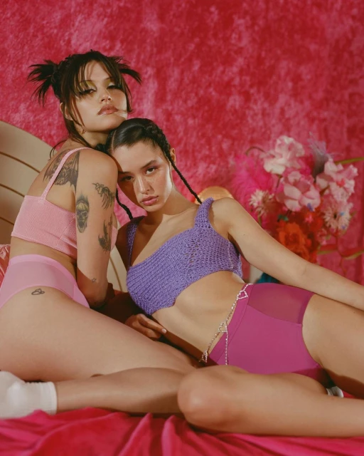 Two young women pose in matching knit tops and bottoms against a pink textured backdrop with floral accents, showcasing fashion.