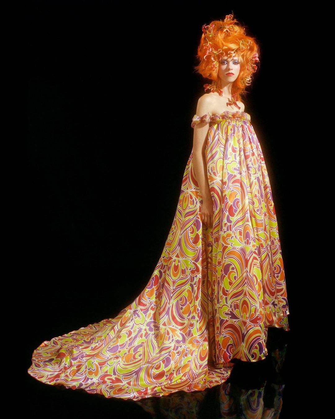 Full-length portrait of a woman in a patterned gown with an orange wig, set against a black background, studio lighting.