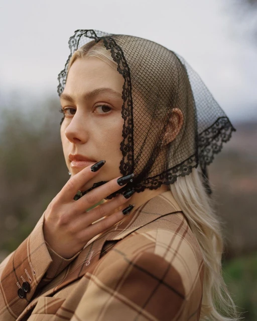 Portrait of a blonde woman wearing a black lace headscarf and a plaid jacket, posing with her hand near her face in an outdoor setting.