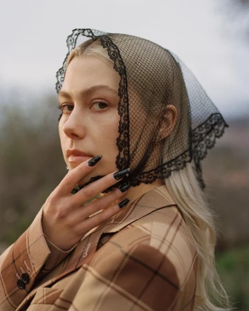 Portrait of a blonde woman wearing a black lace headscarf and a plaid jacket, posing with her hand near her face in an outdoor setting.