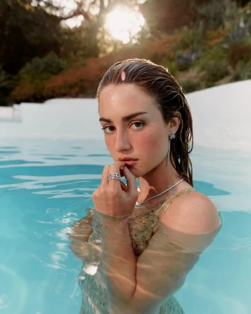 Wet-haired woman standing in an outdoor pool wearing diamond jewellery and a patterned swimsuit under warm sunlight.