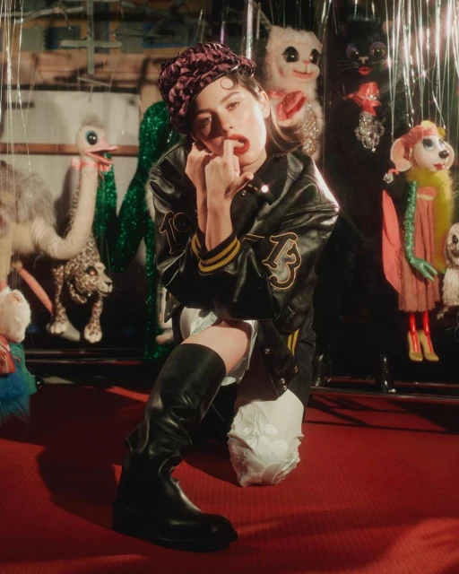 A woman kneels on a red carpet wearing black boots and a jacket. Decorative puppets are in the background, illuminated by soft light.