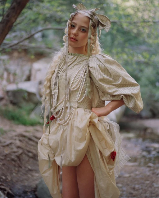 Young woman in a whimsical fashion dress with ribbons and pearls, posing in an outdoor forest-like environment with stone features.