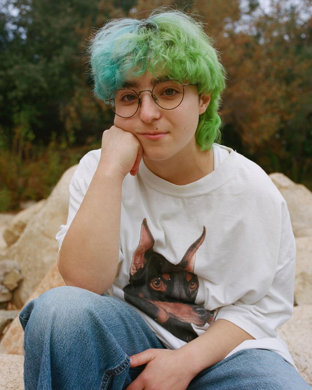 Portrait of a young adult with dyed hair, wearing a dog-graphic tee and jeans, sitting outdoors among rocks and greenery.