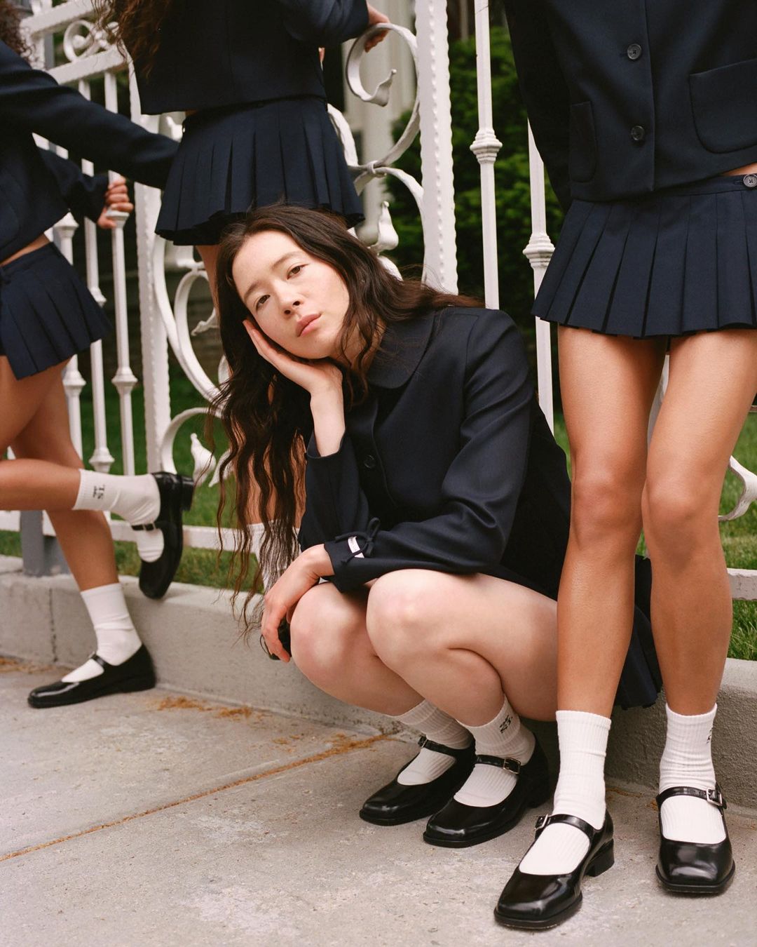 Fashion shot of a young woman crouching, surrounded by three other women in navy skirt suits and white socks outside.