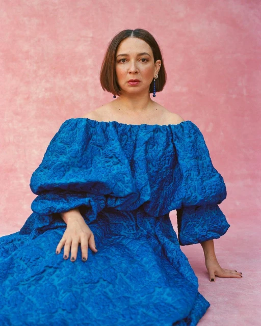 Portrait of adult female with bob haircut in a large blue dress with textured material on a pink textured background, posed.