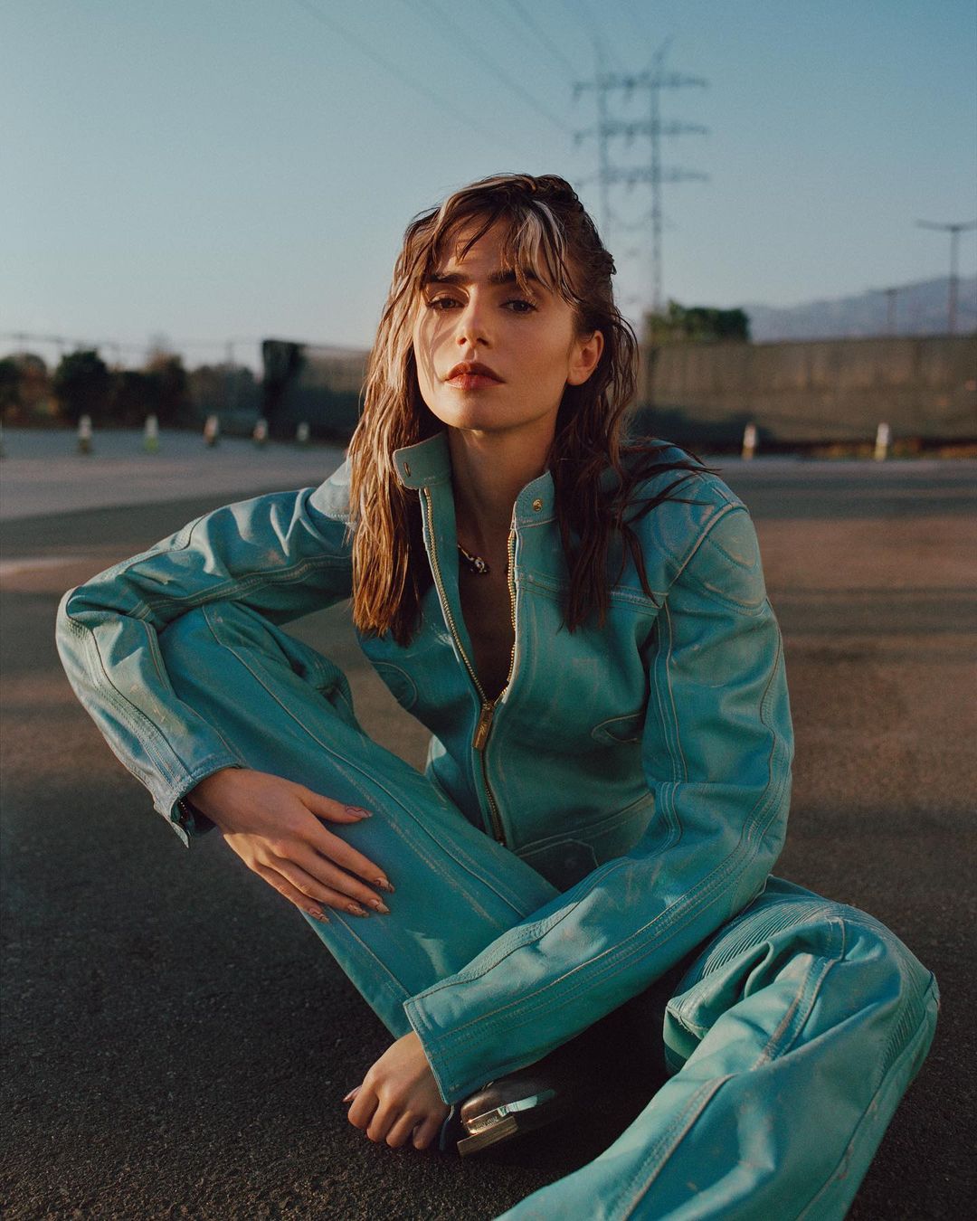 Woman with wet brown hair sits on asphalt in an aquamarine jumpsuit with powerlines and building in background under a clear sky.