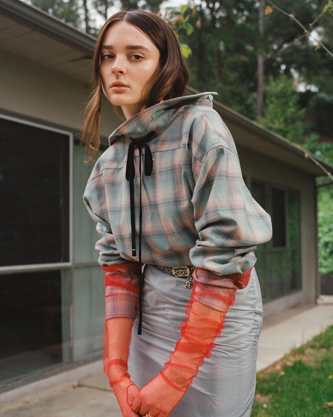 A woman poses in a plaid top and grey skirt against a home exterior backdrop, creating a fashion-forward, outdoor editorial image.