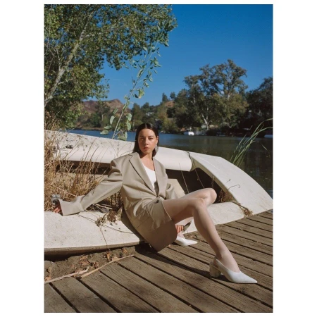 Woman in an oversized tan blazer sitting on a weathered boat on a wooden dock by a calm lake under a clear blue sky