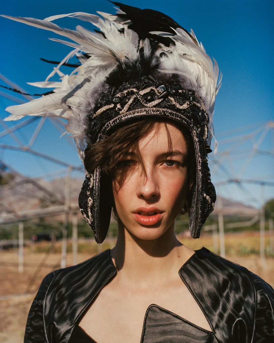 Portrait of a woman wearing a feather headdress and a structured black jacket against an outdoor backdrop with a greenhouse structure.