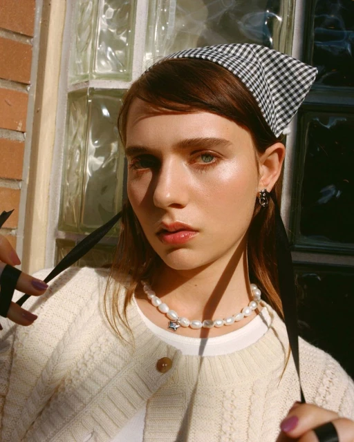 Close-up of a young woman with a gingham headscarf, pearl necklace with a star pendant, wearing a cream-colored sweater. Outdoor setting.