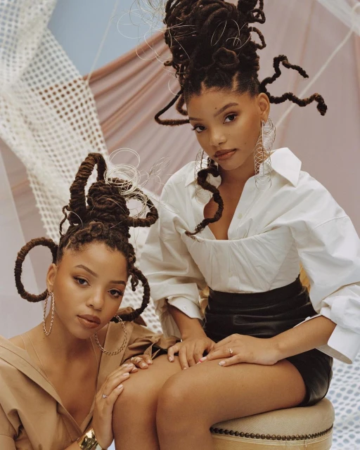 Two Black women pose indoors with creative hair styling. One wears a white shirt and the other a neutral-tone blouse with gold jewelry.
