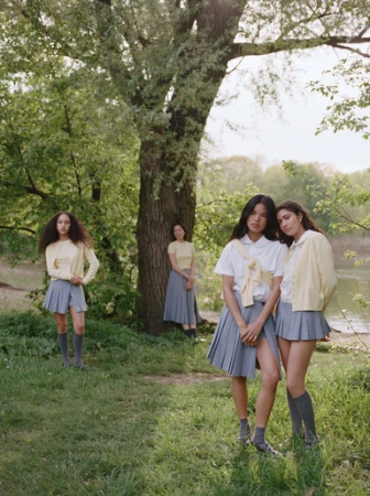 Four women in school uniforms are standing on a grassy field with trees in the background on a sunny day, fashion editorial outdoors.