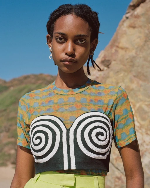 Portrait of a Black woman in a graphic-print top and green pants outdoors, posing near rock formations against a clear blue sky.