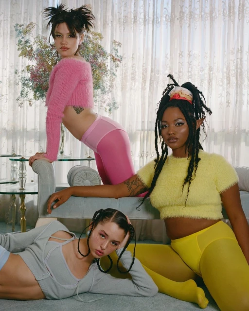 Three diverse female models showcase fashion in an indoor setting, wearing pink, yellow, and gray tops and leggings, with neutral window light.