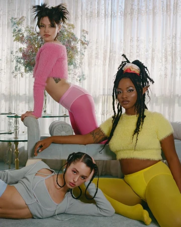 Three diverse female models showcase fashion in an indoor setting, wearing pink, yellow, and gray tops and leggings, with neutral window light.