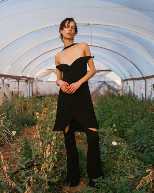 A slender adult in a black dress poses in a greenhouse, bathed in soft natural light, standing amidst green plants and translucent coverings.