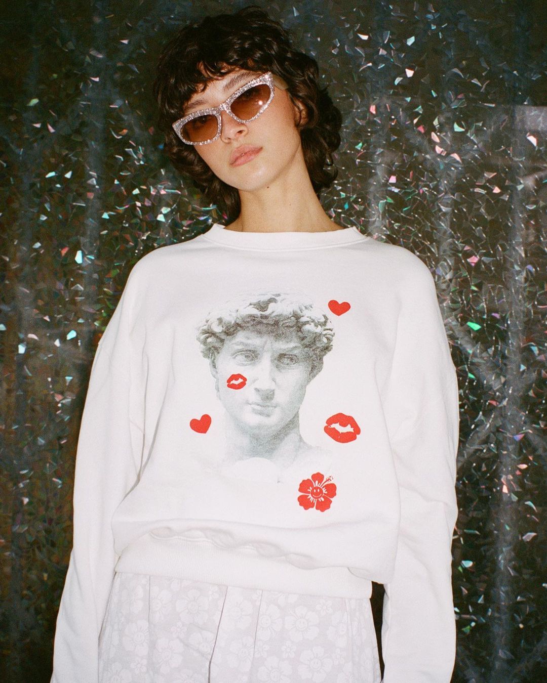 A brunette female wears a white sweater with a classical bust graphic, pink sunglasses, and floral pants against a dark, shiny background.