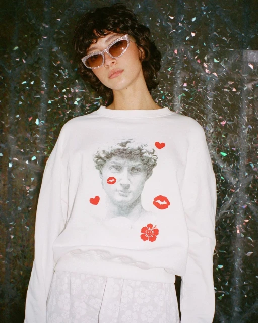A brunette female wears a white sweater with a classical bust graphic, pink sunglasses, and floral pants against a dark, shiny background.