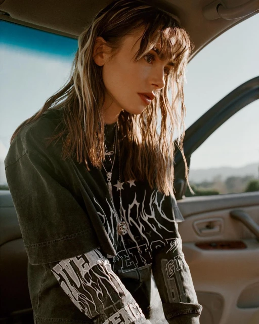 Woman with long hair and graphic tee sitting in a car, looking down. Automotive interior in neutral colors is visible.