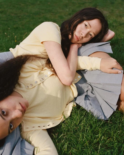 Two young women pose on grass in yellow tops and gray skirts. One woman leans on the other in a relaxed, outdoor setting.