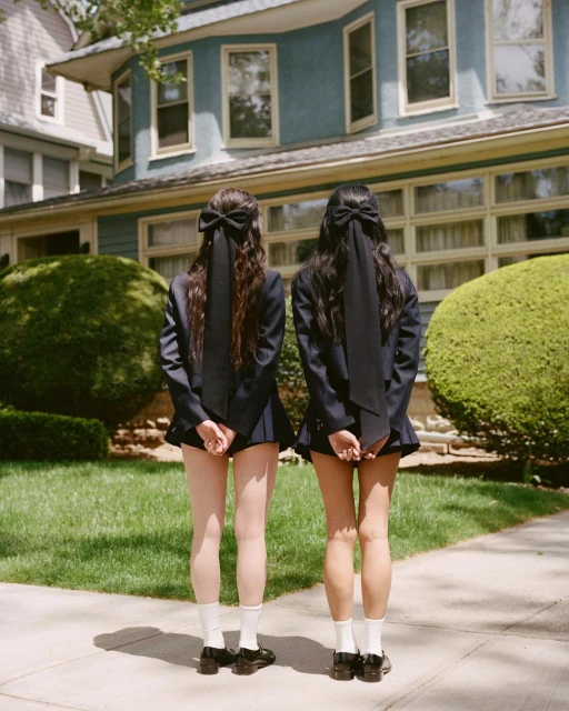 Two young women in dark jackets and skirts with bows in their hair stand on a sidewalk facing a house with green lawn and bushes.