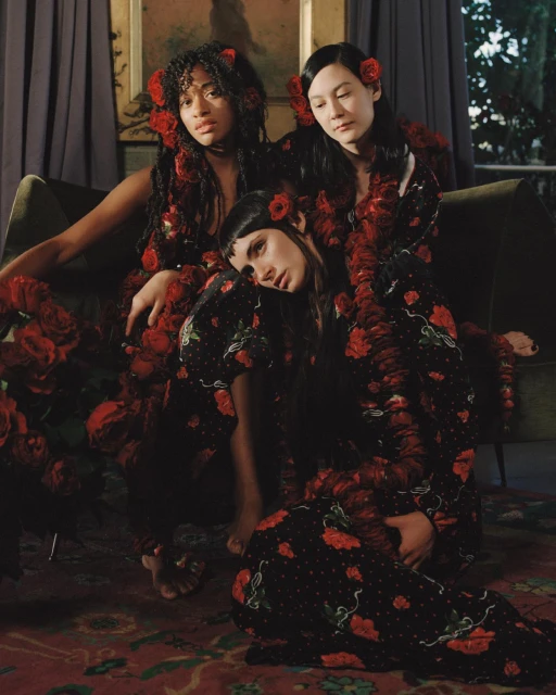 Three women pose in black floral-print dresses with red roses in their hair against a dark indoor backdrop, creating a vintage aesthetic.