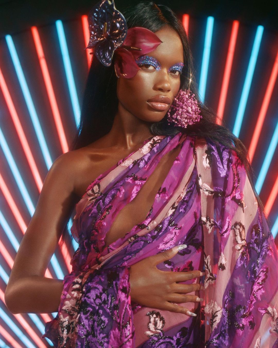 Close-up studio portrait of a dark-skinned woman with glittery makeup, floral hair accessory, and a patterned purple shawl, against blue and red neon lights.