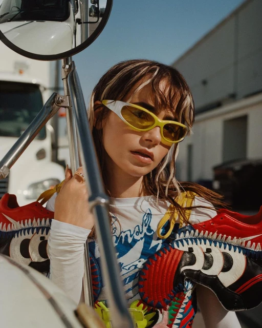 Stylish woman with yellow glasses and brown hair poses in front of a white vehicle, adorned with cartoon-themed accessories and clothing.