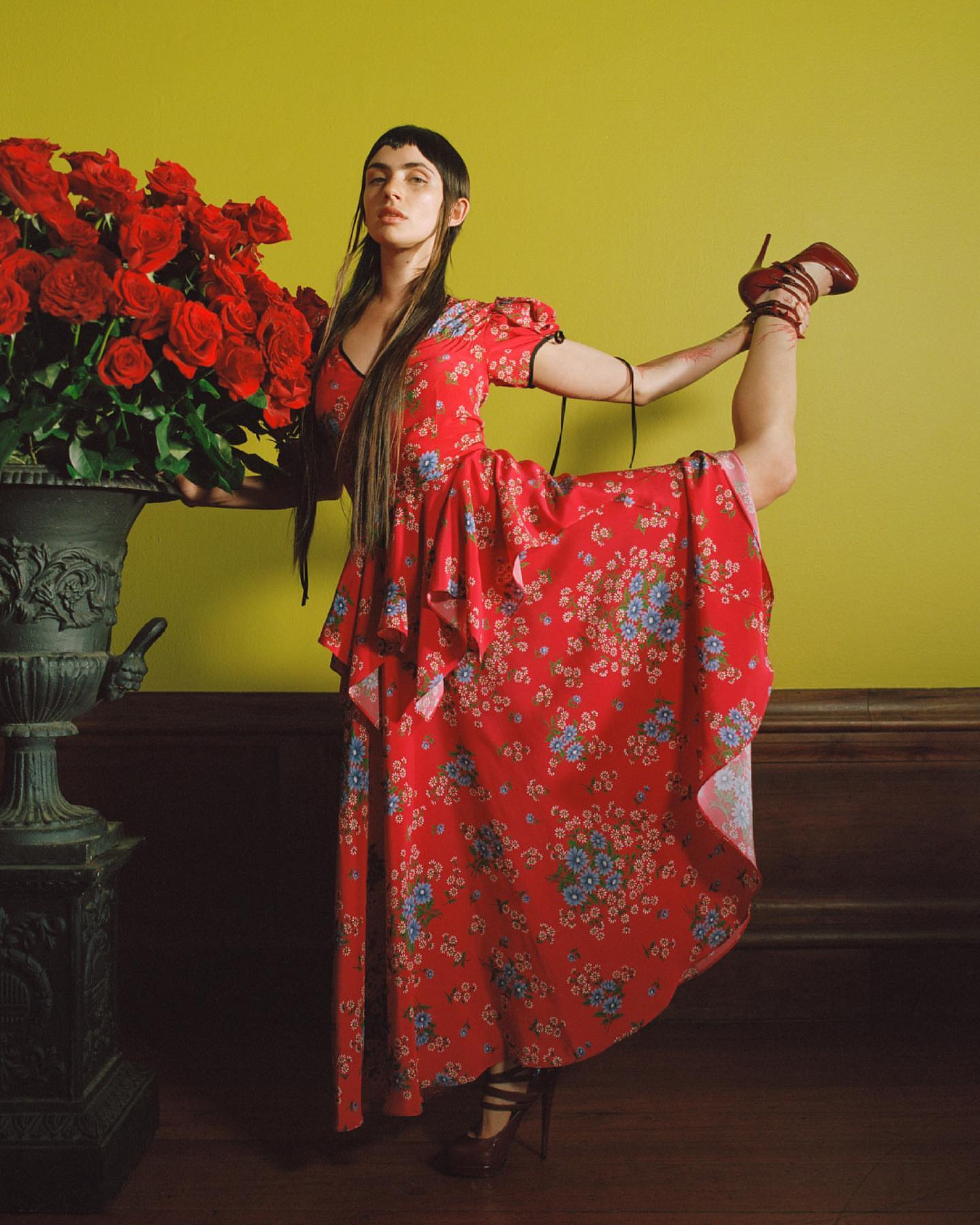 A woman in a red floral dress balances with one leg raised behind her, supported by her hand, against a backdrop of red roses.
