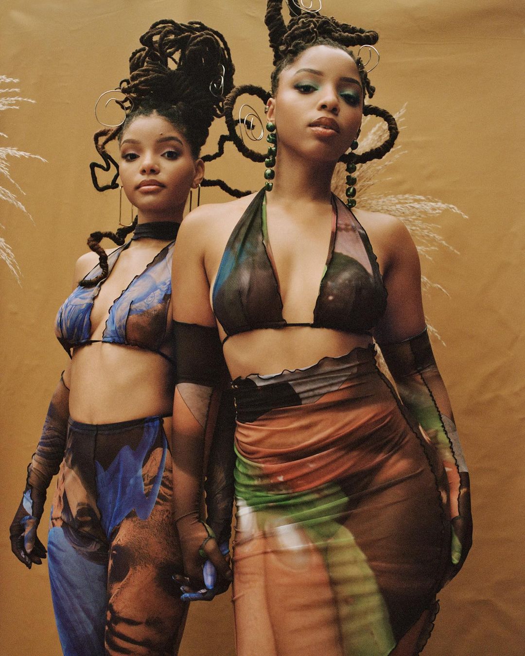 Two women with dreadlocks pose in coordinated outfits against a neutral backdrop, showcasing fashion and style.