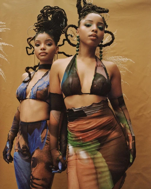 Two women with dreadlocks pose in coordinated outfits against a neutral backdrop, showcasing fashion and style.