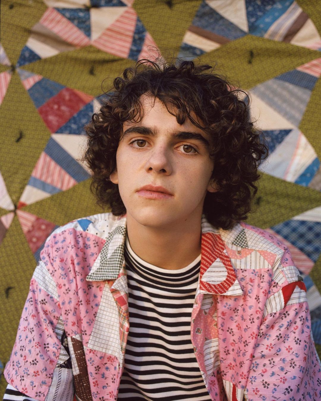 Portrait of a young adult with curly hair, wearing a striped shirt and pink patchwork jacket, posing in front of a quilt.