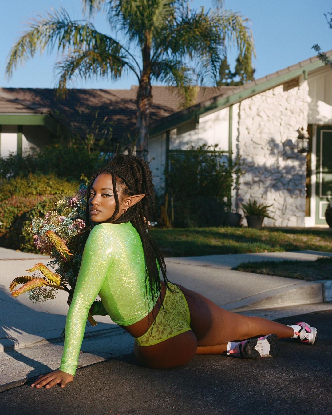 Woman in neon green top and shorts poses on a paved road with a house and palm trees in the background on a sunny day.