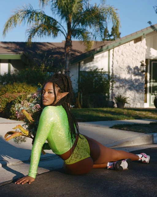 Woman in neon green top and shorts poses on a paved road with a house and palm trees in the background on a sunny day.